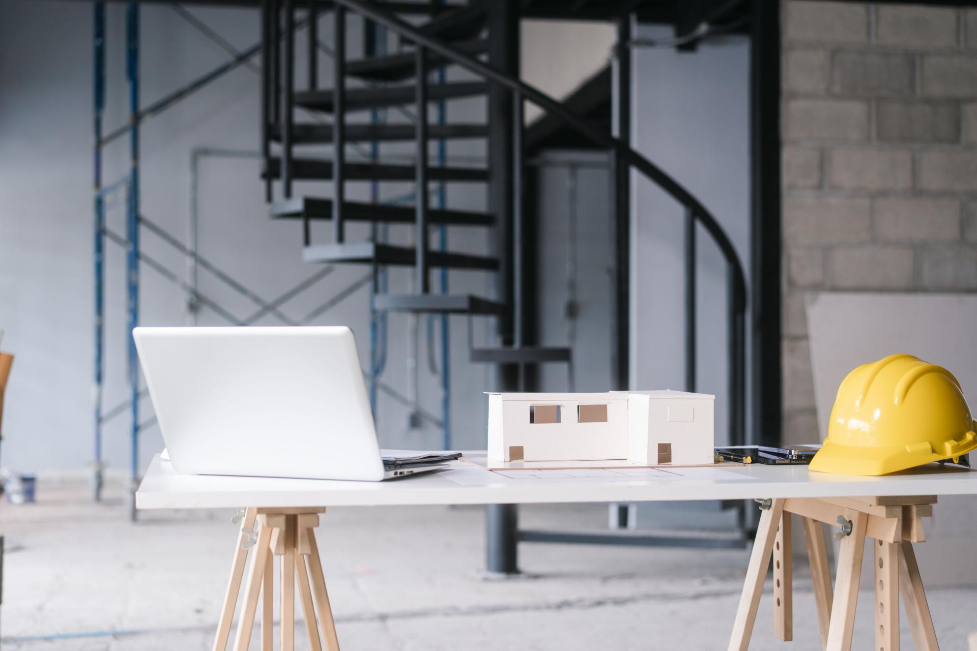 Modern architectural workspace featuring white laptop, scale house model, yellow safety helmet on table. Spiral staircase and scaffolding in background suggest active construction and design planning